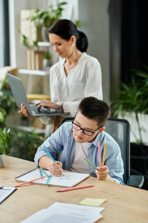 A young boy with Down syndrome draws at a table while his mother works on a laptop in the background.の写真素材