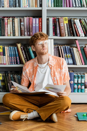 A man sits on the floor in front of a bookshelf.