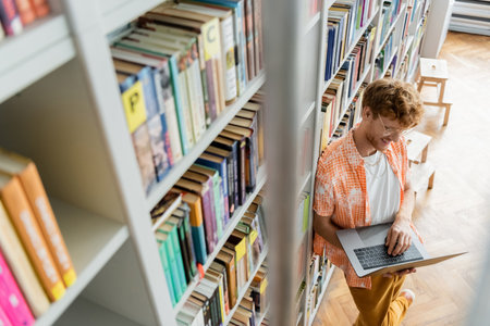 A young man deeply focused on his laptop amidst stacks of books in a library.