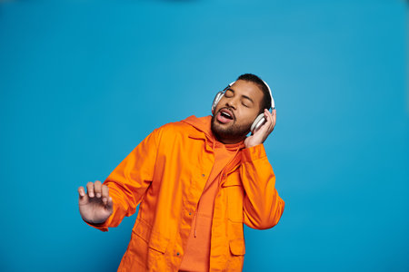 african american man in orange outfit and headphones singing with closed eyes on blue background