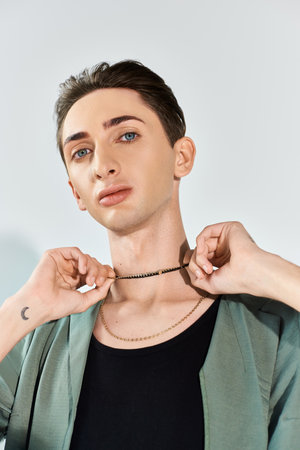 A young queer man adjusts his necklace, showcasing pride and elegance in a studio setting against a grey background.の写真素材