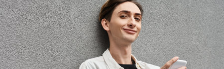 A stylishly dressed young queer person leans against a wall, engrossed in his cell phone.
