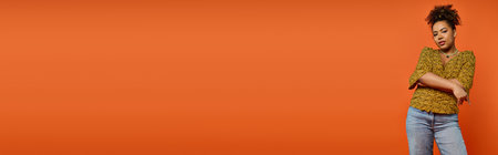 African American woman in stylish attire, arms crossed, on vibrant orange backdrop.