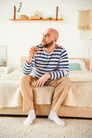 A stylish man in glasses sits atop a bed in a modern bedroom, contemplating the day.の写真素材
