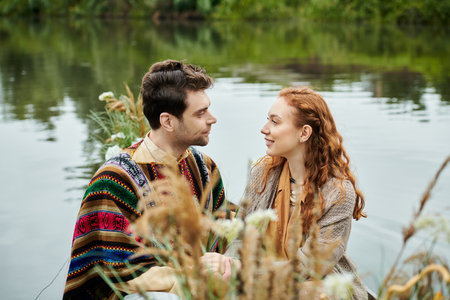 A man and woman in boho attire relax by a serene body of water, enjoying a romantic date in a lush green park setting.の写真素材