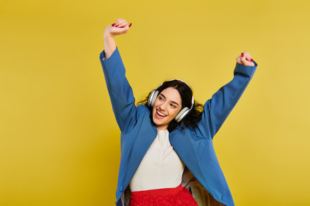Brunette woman with curly hair poses confidently, raising her hands in a blue jacket against a vibrant yellow studio backdrop.の写真素材