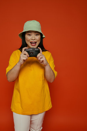 An Asian woman in a yellow shirt captures moments with a camera on in a studio on an orange...