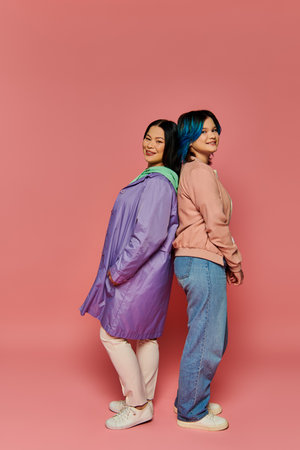 An Asian mother and her teenage daughter stand together in a casual pose against a vibrant pink...