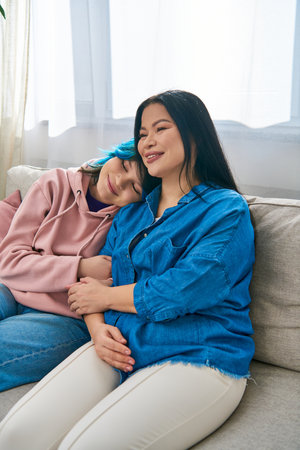 An Asian mother and her teenage daughter in casual wear sit together on top of a couch, sharing a...