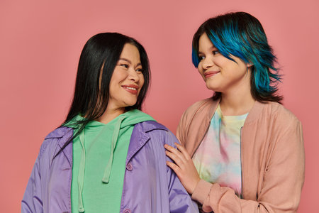 Asian mother and teenage daughter standing together in casual wear in front of a vibrant pink...