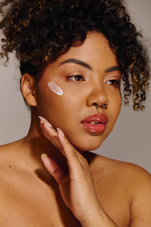 A beautiful young African American woman with curly hair showcasing a white cream on her cheek in a studio setting.の写真素材
