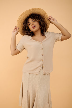 A stunning young African American woman, with curly hair, elegantly poses in a summer dress and stylish hat in a studio setting.の写真素材