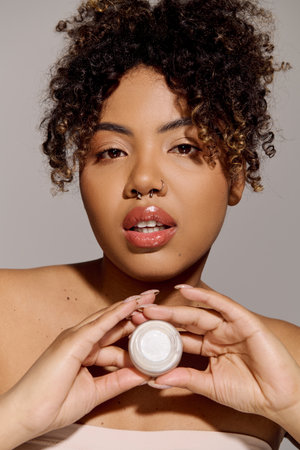 A beautiful African American woman with curly hair holding a jar of cream delicately in her hands.