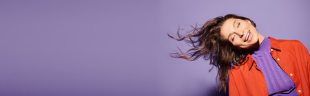 A stylish young woman in vibrant orange attire stands against a purple background, her hair gracefully blowing in the wind.の写真素材