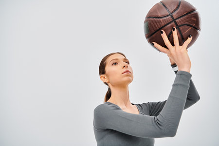 A sporty young woman in active wear holding a basketball up in the air against a grey background.の写真素材