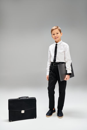 A stylish young boy stands confidently next to a briefcase on a gray backdrop.の写真素材