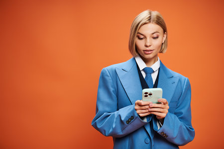 positive young woman with blonde short hair posing with her smartphone on orange background