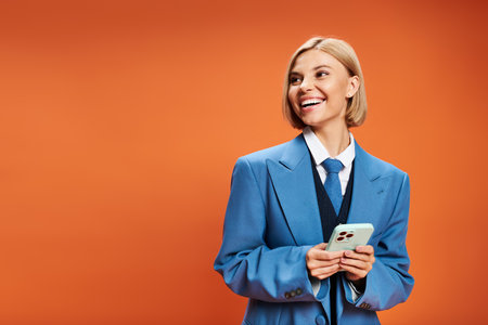 joyful well dressed woman with short blonde hair in chic attire holding phone on orange backdropの写真素材
