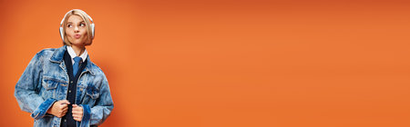happy young woman with blonde hair with headphones in denim jacket posing on orange backdrop, bannerの写真素材
