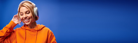merry attractive woman with short blonde hair and headphones enjoying music on blue backdrop, bannerの写真素材