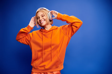 good looking stylish woman with short blonde hair and headphones enjoying music on blue backdropの写真素材