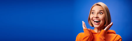 positive woman with short hair in vibrant orange hoodie posing actively on blue backdrop, bannerの写真素材