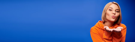 cheerful woman with short hair in vibrant orange hoodie posing actively on blue backdrop, bannerの写真素材