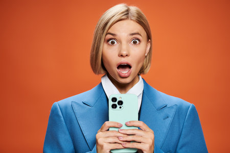 surprised young woman with blonde short hair posing with her smartphone on orange backgroundの写真素材