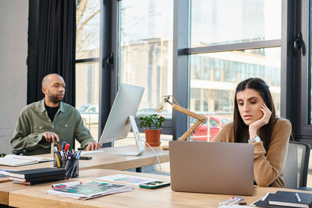 A man and a woman sit at a table in an office, focused on a laptop screen, working together on a...