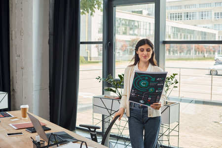 A businesswoman stands confidently in front of a window, holding charts with a thoughtful...