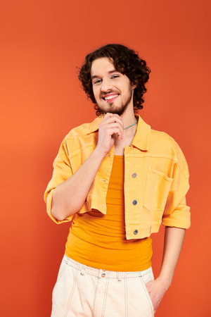 contented appealing gay man with dark hair and vibrant makeup posing on orange backdrop, pride monthの写真素材