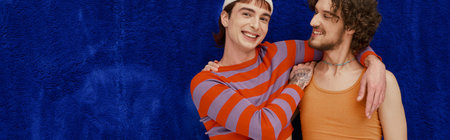 two merry good looking gay men in vibrant clothes posing on dark blue backdrop, pride month, banner