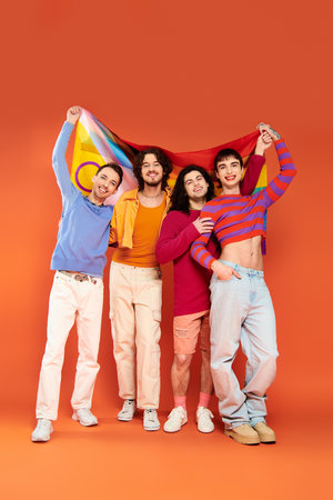 four joyful fashionable gay men in vibrant attires holding rainbow flag in front of camera, prideの写真素材