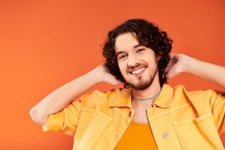joyous appealing gay man with dark hair and vibrant makeup posing on orange backdrop, pride month