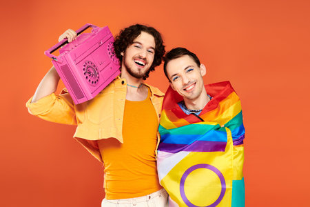 two cheerful handsome gay friends posing with tape recorder and rainbow flag on orange backdropの写真素材
