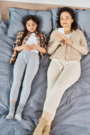 A mother and daughter are laying on a bed, absorbed in their smartphones, enjoying quality time together at home.の写真素材