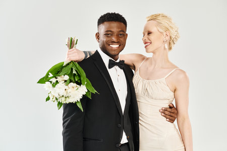 A beautiful blonde bride in a wedding dress and an African American groom in a tuxedo standing confidently together in a studio.の写真素材