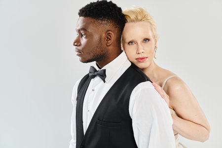 A beautiful blonde bride in a wedding dress standing next to an African American groom in a tuxedo against a grey studio backdrop.の写真素材