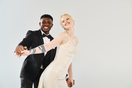 A beautiful blonde bride in a wedding dress stands beside an African American groom in a sharp tuxedo on a grey background.の写真素材