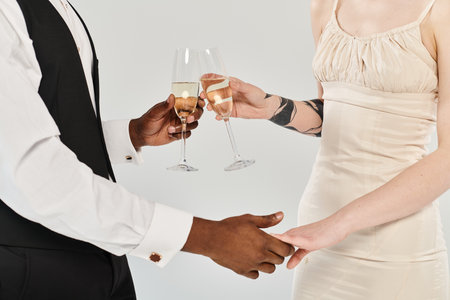 A beautiful blonde bride in a wedding dress and an African American groom holding glasses of champagne in a studio on a grey background.の写真素材