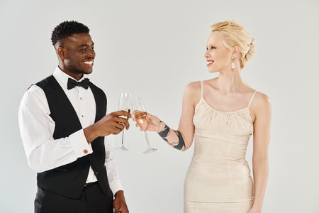 Beautiful blonde bride in wedding dress and African American groom holding champagne glasses in a studio on a grey background.の写真素材