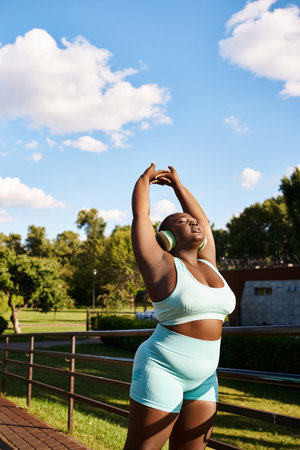 An African American woman in a blue top and shorts energetically stretches her arms outdoors, embodying body positivity.の写真素材