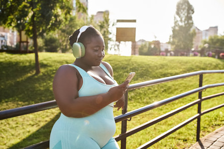 An African American woman, embracing her body positivity, wearing headphones looks intently at her cell phone while enjoying music outdoors.の写真素材