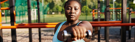 A curvy African American woman in a blue sportswear confidently holds dumbbell in her hands, exuding...