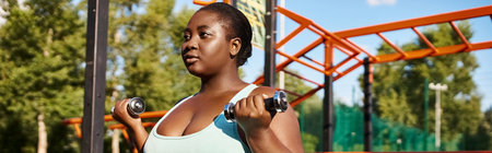 An African American woman in sportswear joyfully lifts dumbbells in front of a colorful playground.の写真素材