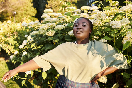 A plus-size African American woman stands confidently in front of a bush of vibrant flowers, embodying body positivity and embracing the beauty of nature.の写真素材