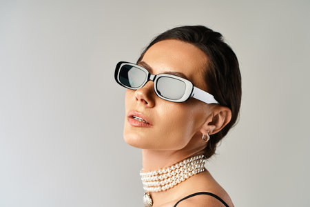 A stylish woman with sunglasses and a pearl necklace exudes confidence in a studio against a grey background.の写真素材