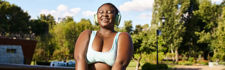 A curvy African American woman in a top standing confidently in a park, listening to music through headphones and embracing her body positivity.の写真素材