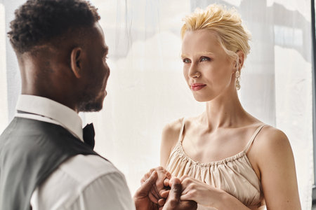 A beautiful blonde bride in a wedding dress and an African American groom stand side by side in a studio against a grey background.の写真素材