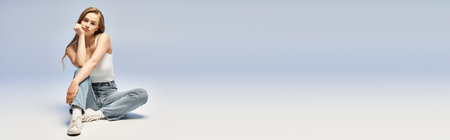 A beautiful young blonde woman gracefully sits on the ground, striking a pose for a photo shoot in a studio setting.の写真素材